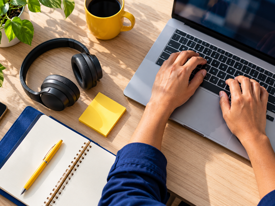 Overhead view of hands typing on a laptop, next to headphones, a yellow sticky note, a yellow pen and an open notebook