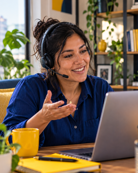 Smiling woman wearing a headset and gesturing during a video call at her laptop, with a yellow mug and a yellow notebook on the desk