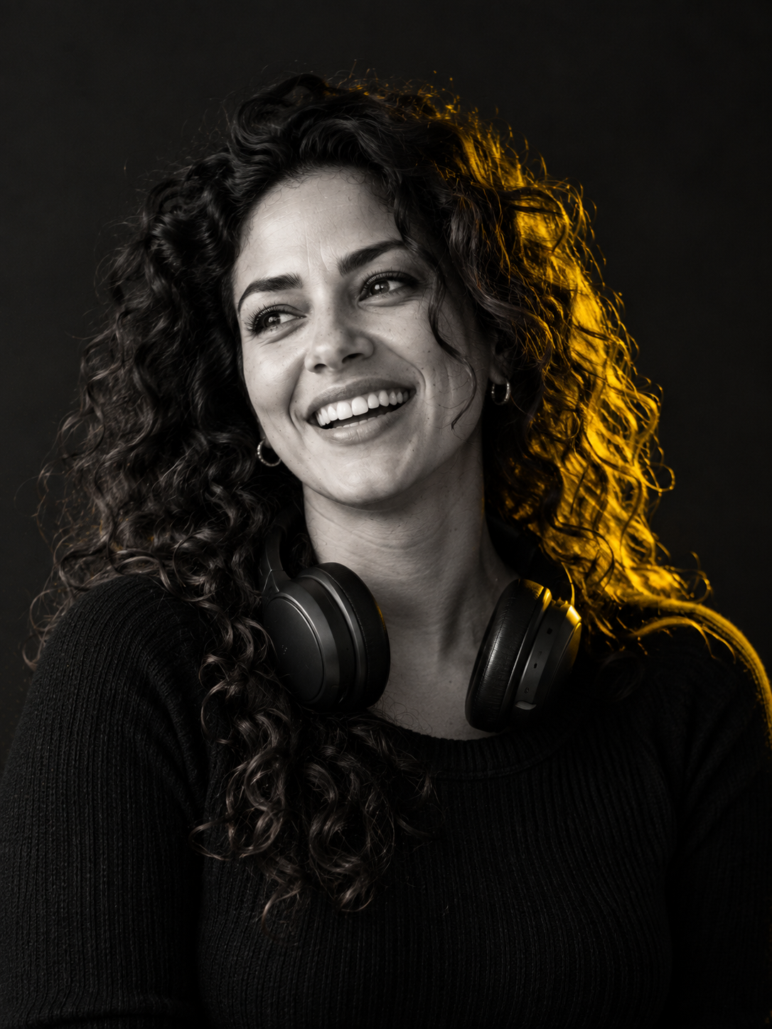 Smiling woman with long curly hair and headphones around her neck, black-and-white portrait with yellow light