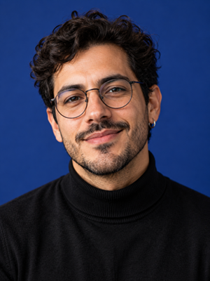 Man with glasses, dark curly hair and a goatee, wearing a black turtleneck against a blue background